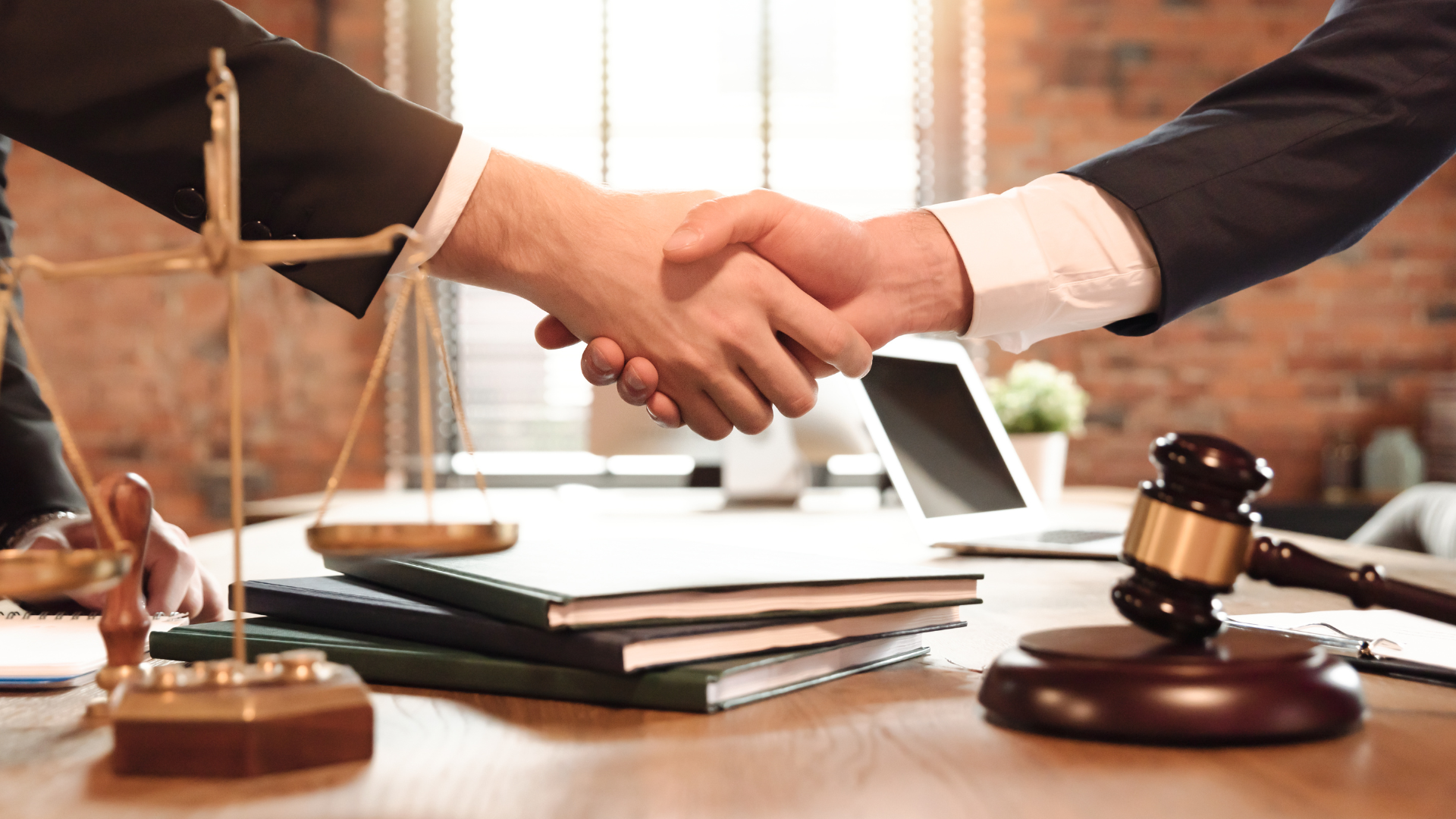 Two people shaking hands over a table with scales, a gavel, and legal documents.