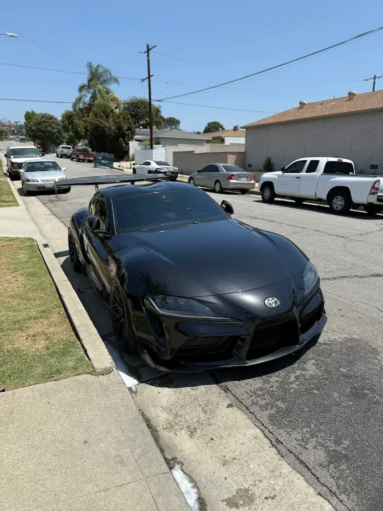 Black sports car parked along a residential curb on a sunny street, with houses and parked vehicles nearby.