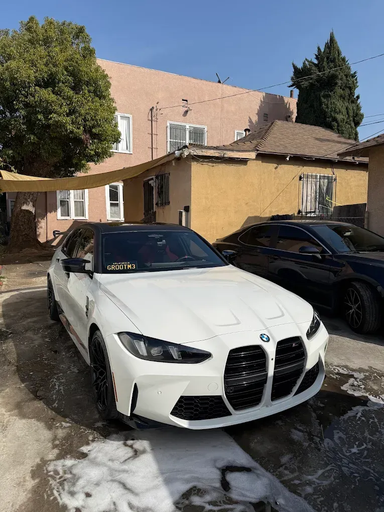 White BMW parked in front of a tan house on a snowy driveway, with another dark car beside it