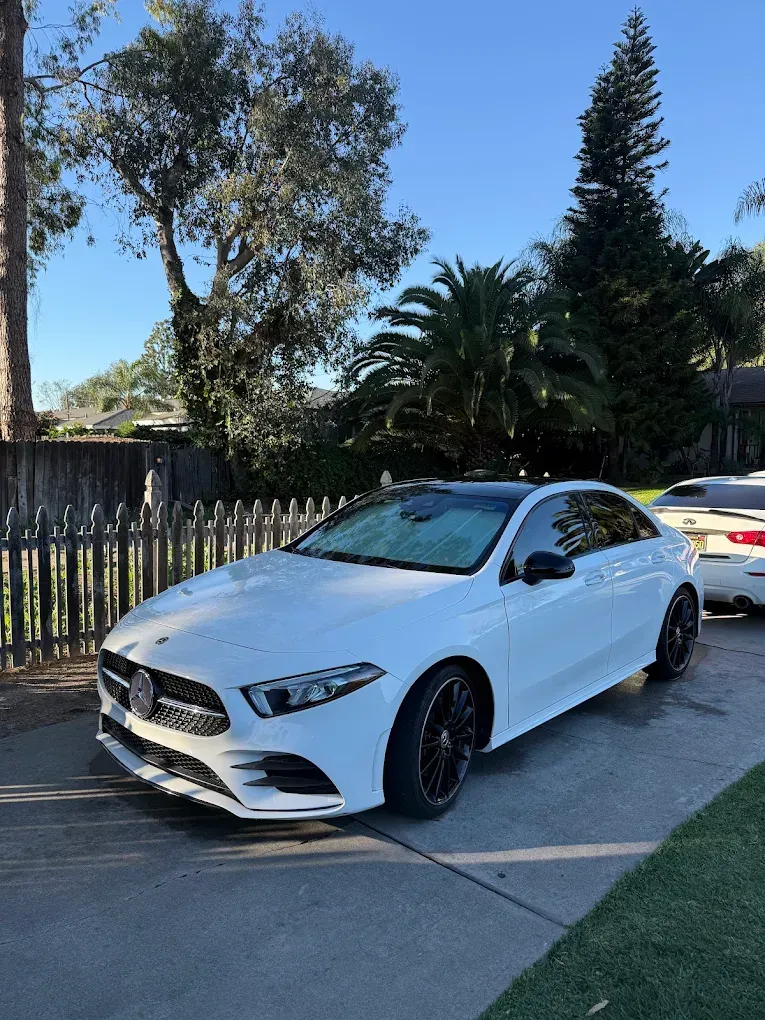White sports sedan parked on a driveway beside a fence and trees on a sunny day