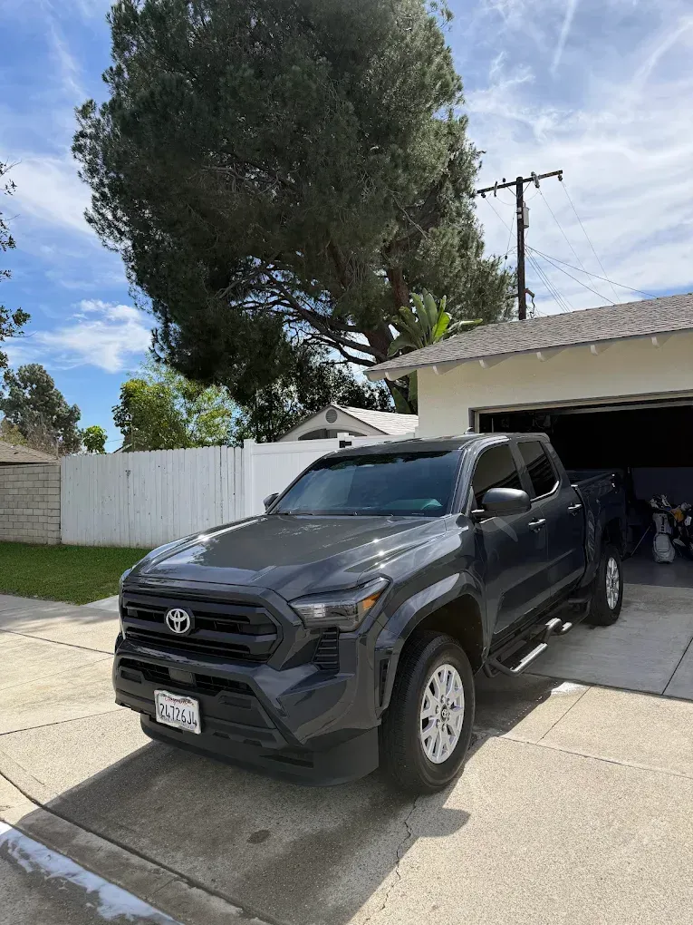 Dark gray pickup truck parked in a driveway beside a garage, with a large tree and white fence behind it