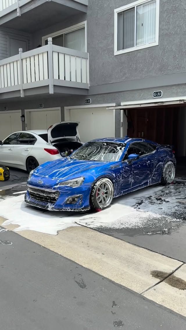 Blue sports car parked in snowy driveway outside a gray townhouse, with a white SUV nearby
