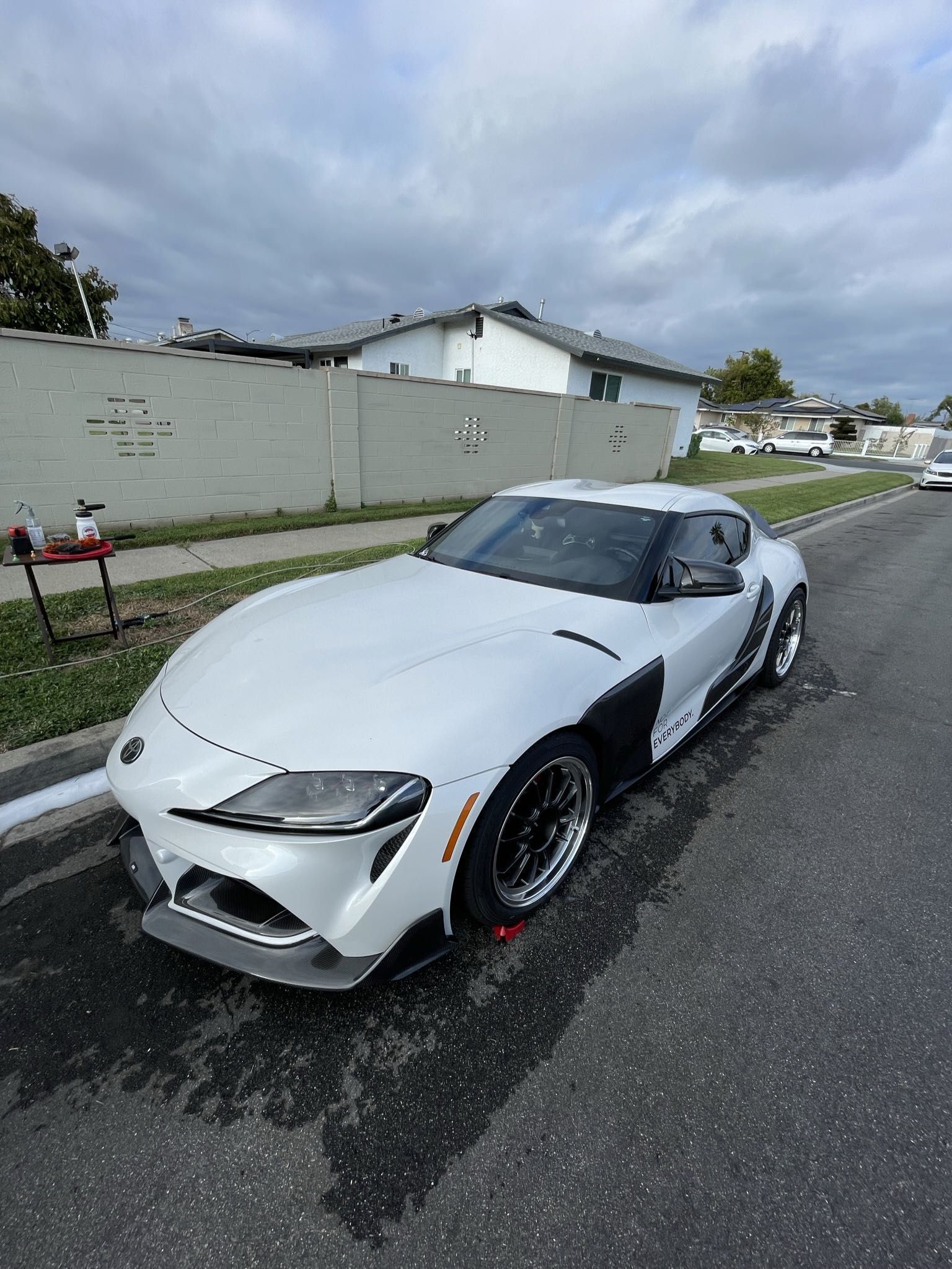 White sports car parked on a wet street beside a gray wall under a cloudy sky