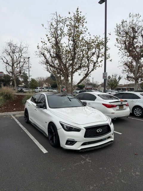 White Infiniti sedan parked in a lot beside other cars and bare trees on an overcast day