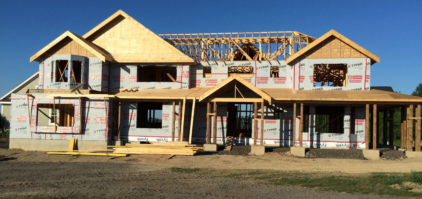 House under construction; wooden frame, tarpaper, blue sky, and grassy foreground.