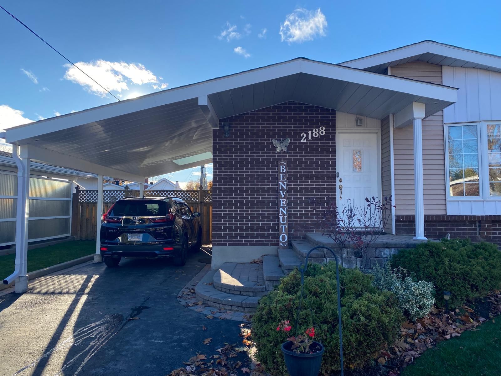 Black SUV parked under a white carport in front of a brick home with a white door. Sunny day.