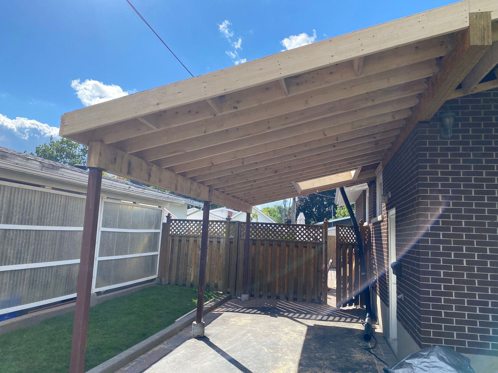 Wooden carport attached to a brick building, over a driveway, with a fence in the background.