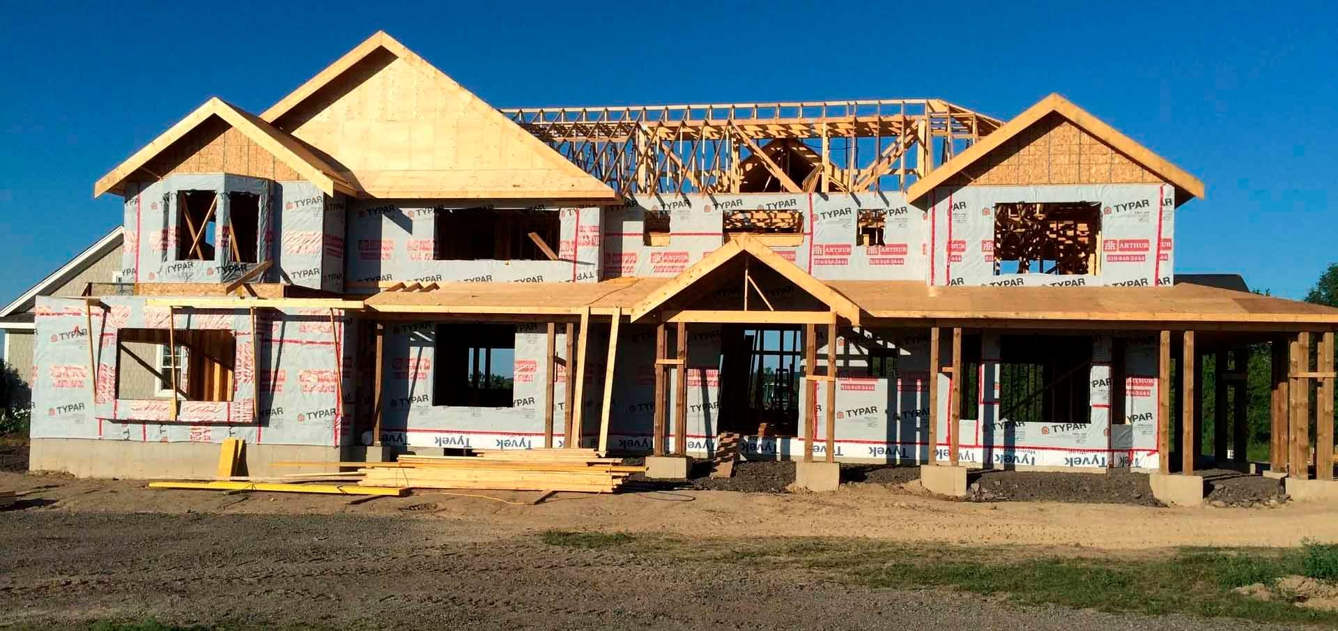 House under construction; wooden frame, tarpaper, blue sky, and grassy foreground.