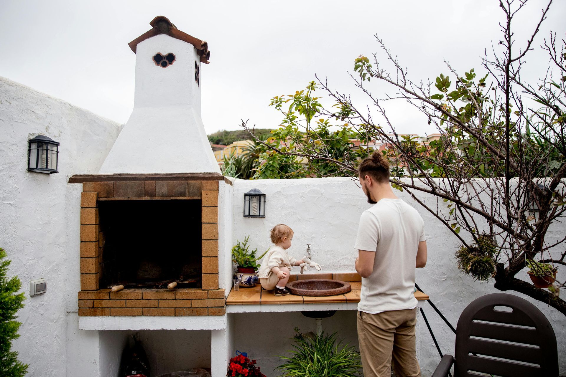 An adult and a small child standing by an outdoor brick fireplace and sink in a white-walled patio area.