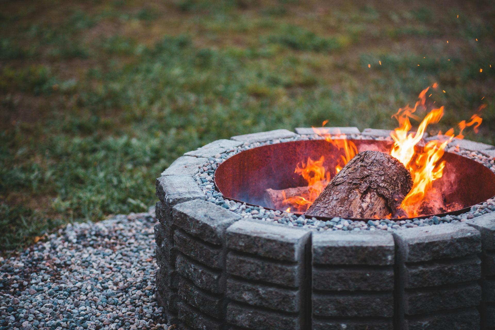 A circular stone fire pit burning with logs on a gravel patio with grass in the background.