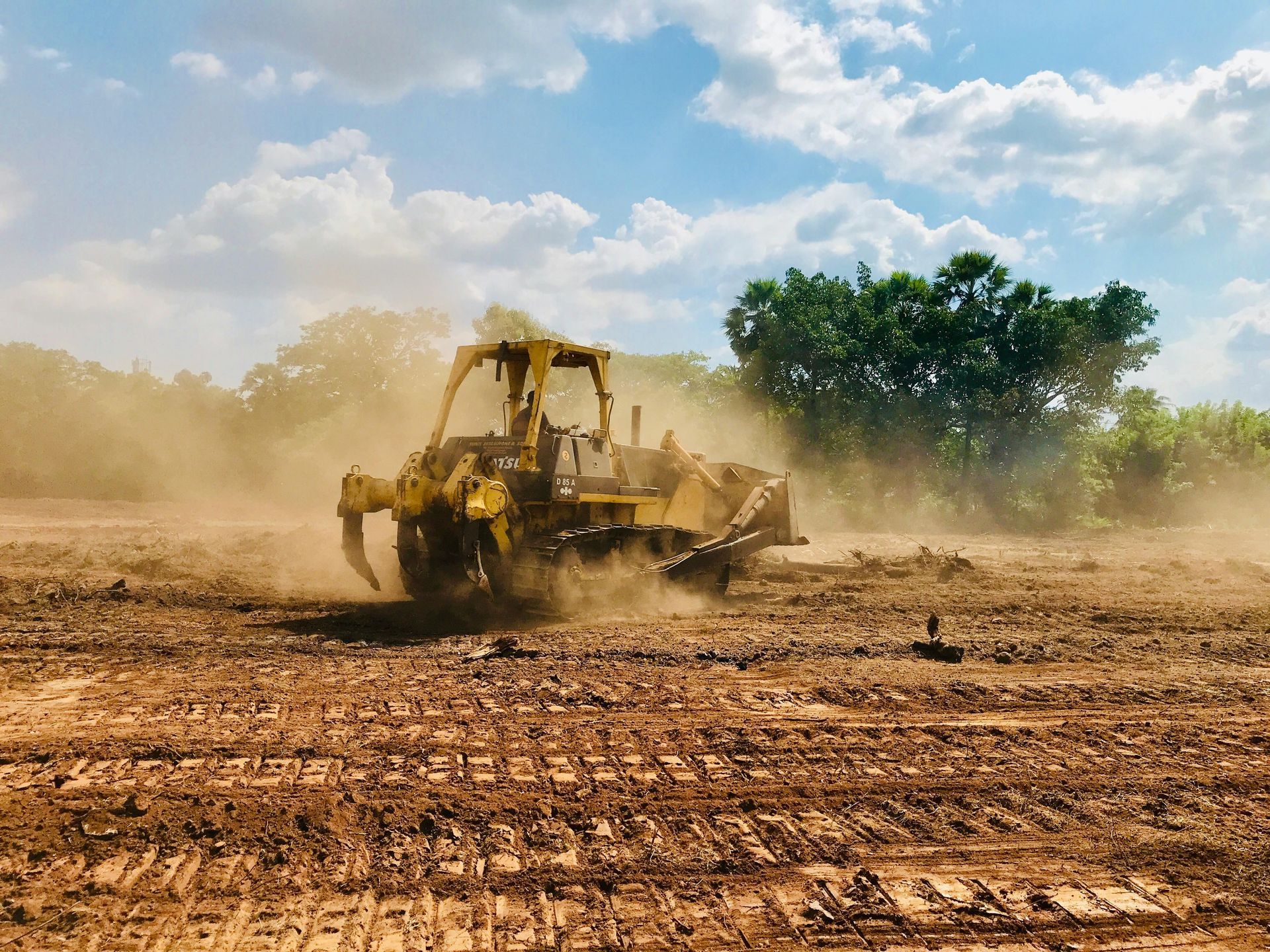 A yellow bulldozer moves across a dusty, churned-up construction site under a bright, cloudy sky.