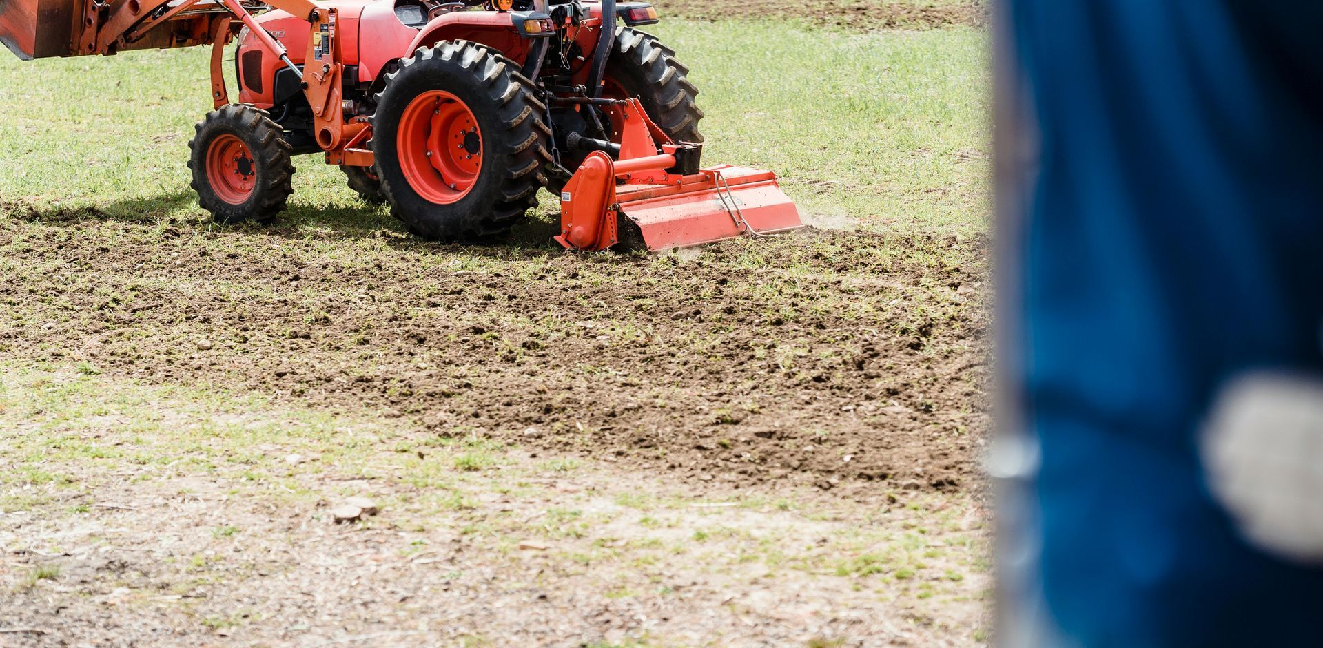 An orange tractor with a tiller attachment turns the soil in a field.