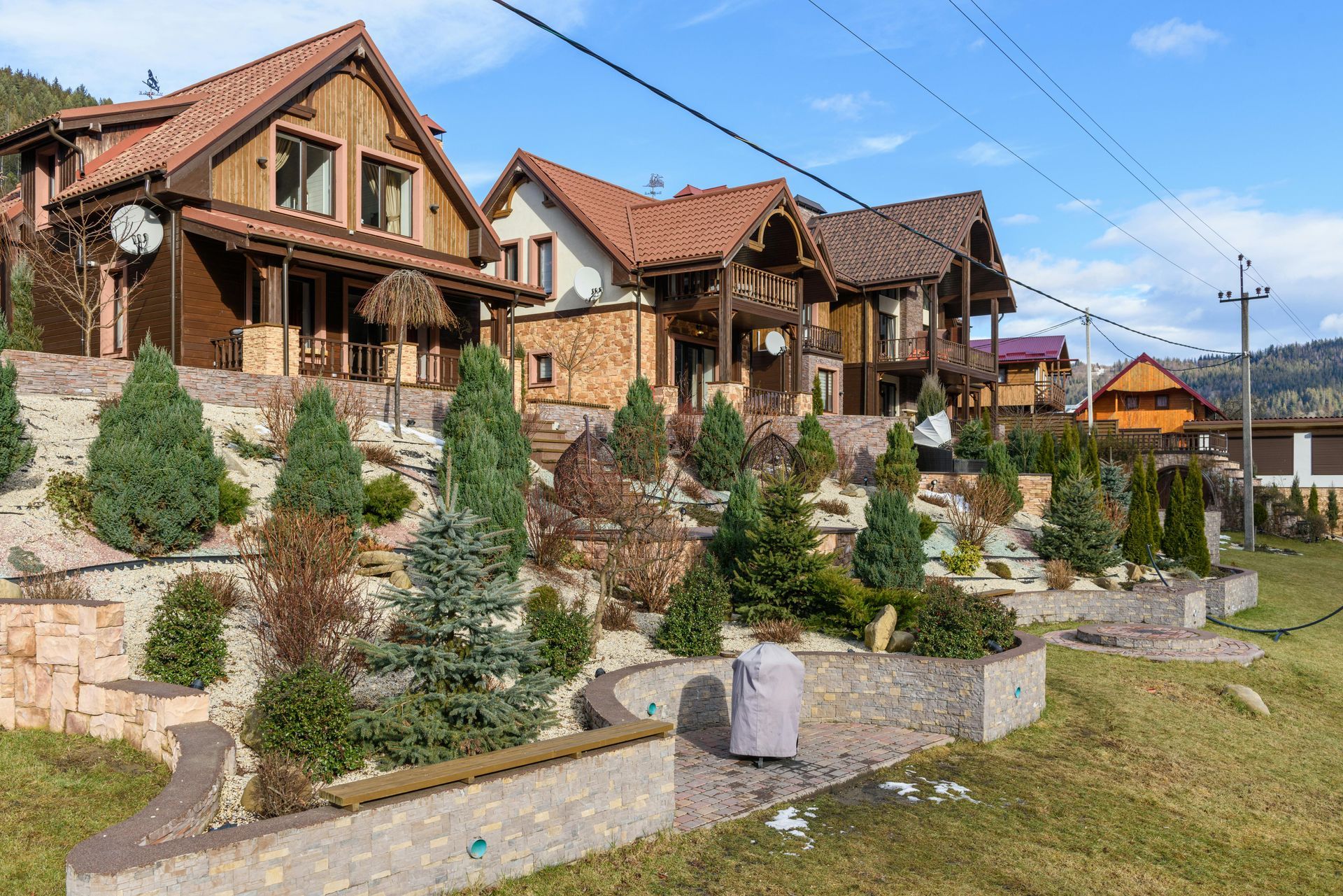 Two multi-story wooden houses with brown tile roofs sit on a terraced, landscaped hill filled with various shrubs and trees.