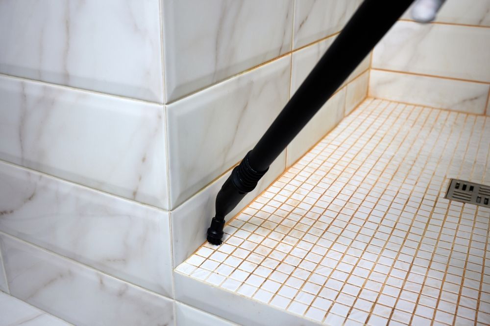 Black steam cleaner nozzle cleaning a shower floor with white and tan tile.