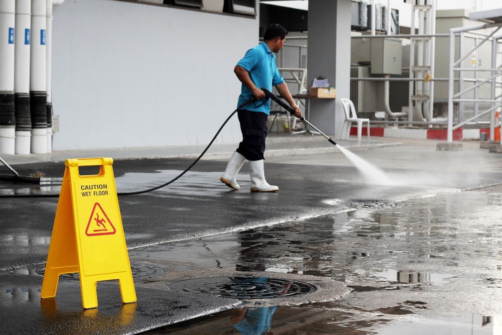 Man in blue shirt power-washing a wet concrete surface, with a caution sign present.