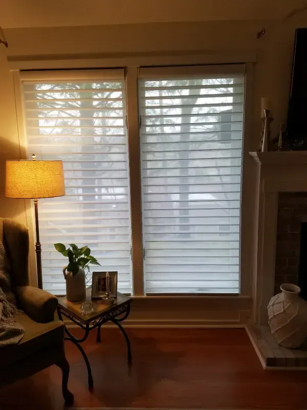 A bright room with two large windows covered by white, horizontal light-filtering shades, a lamp, and a small table.