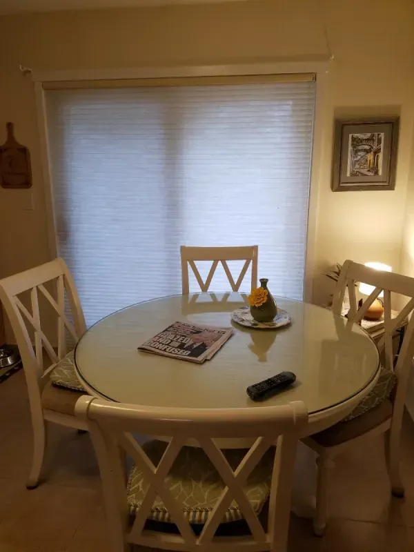 A round white dining table with four matching wooden chairs, a newspaper, and a vase, set before a large window blind.