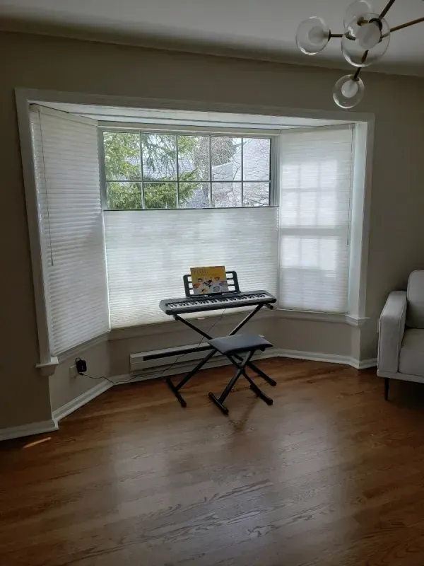 A keyboard on a stand with a music book sits in front of a bay window with white blinds in a room with hardwood floors.