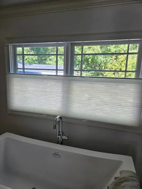 A white bathtub sits below a window featuring a white pleated shade that covers the lower half of the glass.