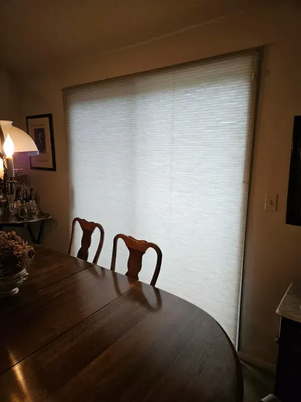 A dining room with a wooden table and two chairs in front of a sliding glass door covered by a white pleated blind.