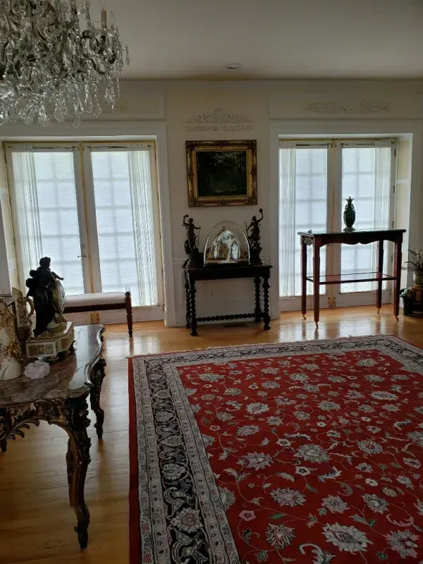 A formal room with a red patterned rug, wood floors, a crystal chandelier, two windows, and decorative furniture.