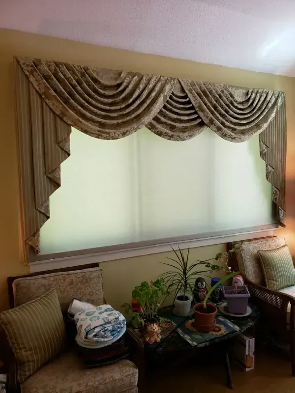 An ornate, pleated tan fabric swag valance hangs over a wide window with light-filtering shades in a sunlit room.