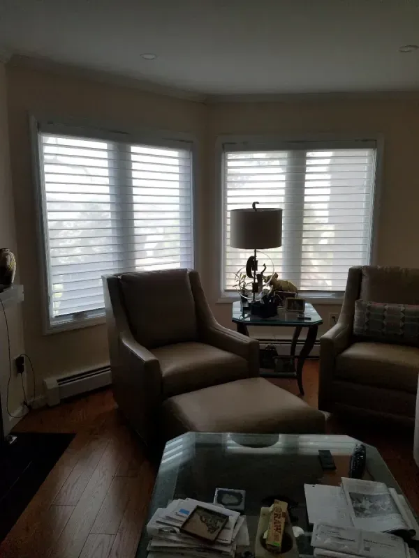 A seating area with two tan armchairs, an ottoman, and a side table with a lamp in front of large windows with blinds.