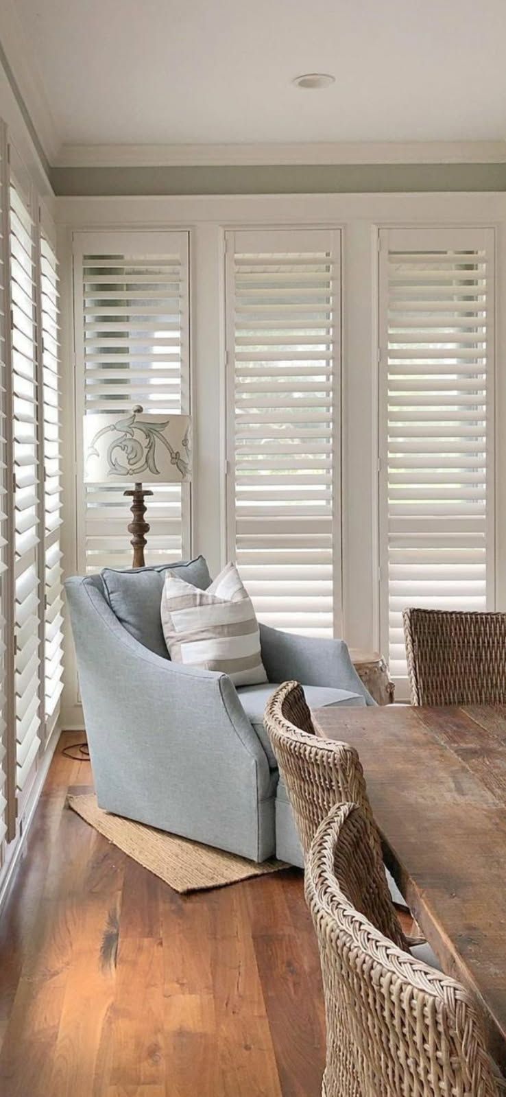 A light blue armchair with a striped pillow sits by a window with white shutters next to a dining table with wicker chairs.