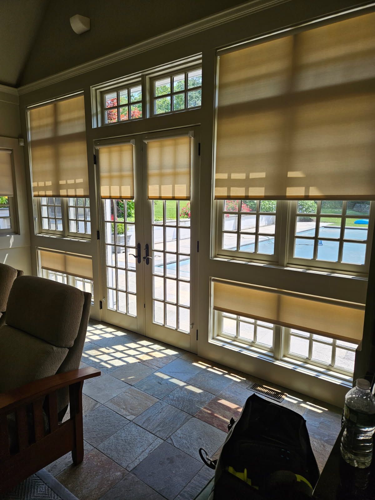 A bright sunroom features a set of French doors flanked by large windows with tan roller shades, overlooking a backyard.
