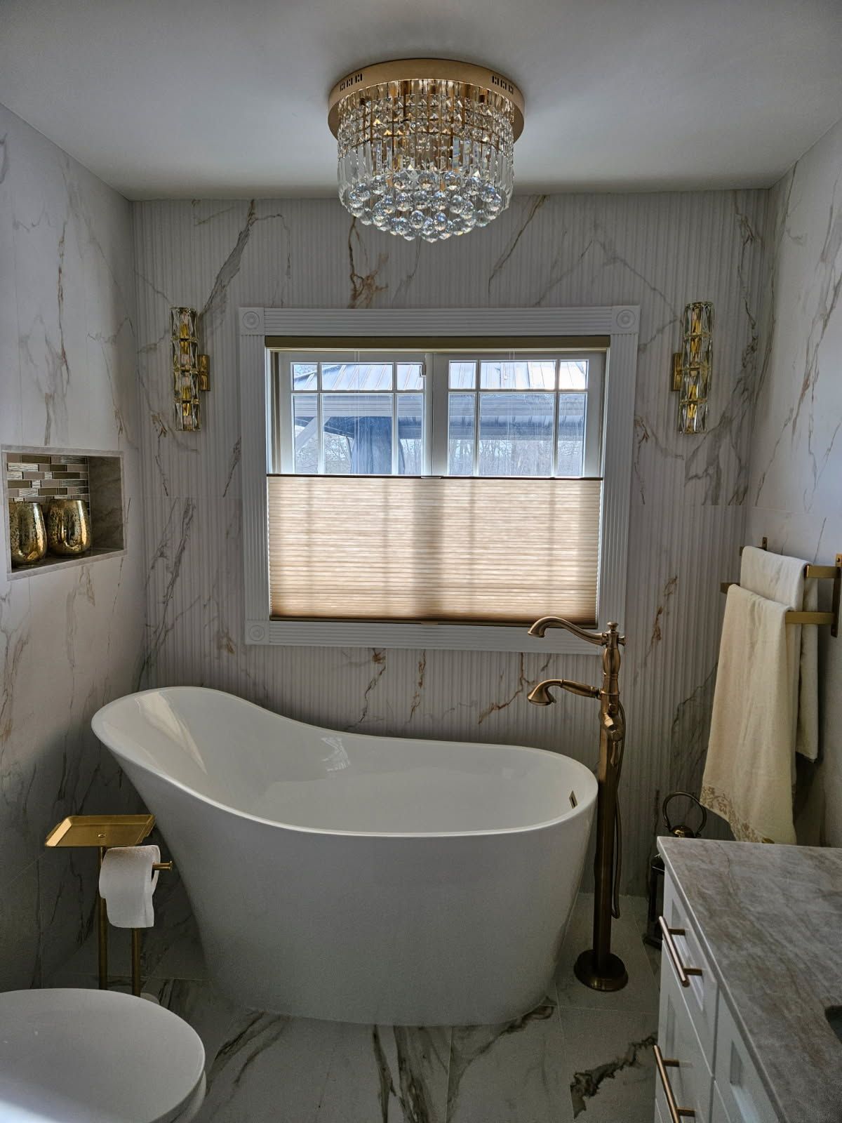 A luxurious bathroom with a white freestanding tub, gold fixtures, marble walls, and a crystal chandelier.
