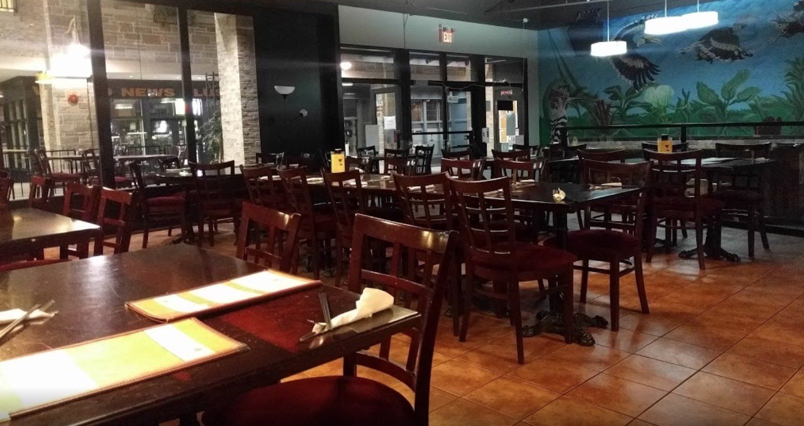 Restaurant interior with dark wood tables and chairs, empty, with large windows and a mural.