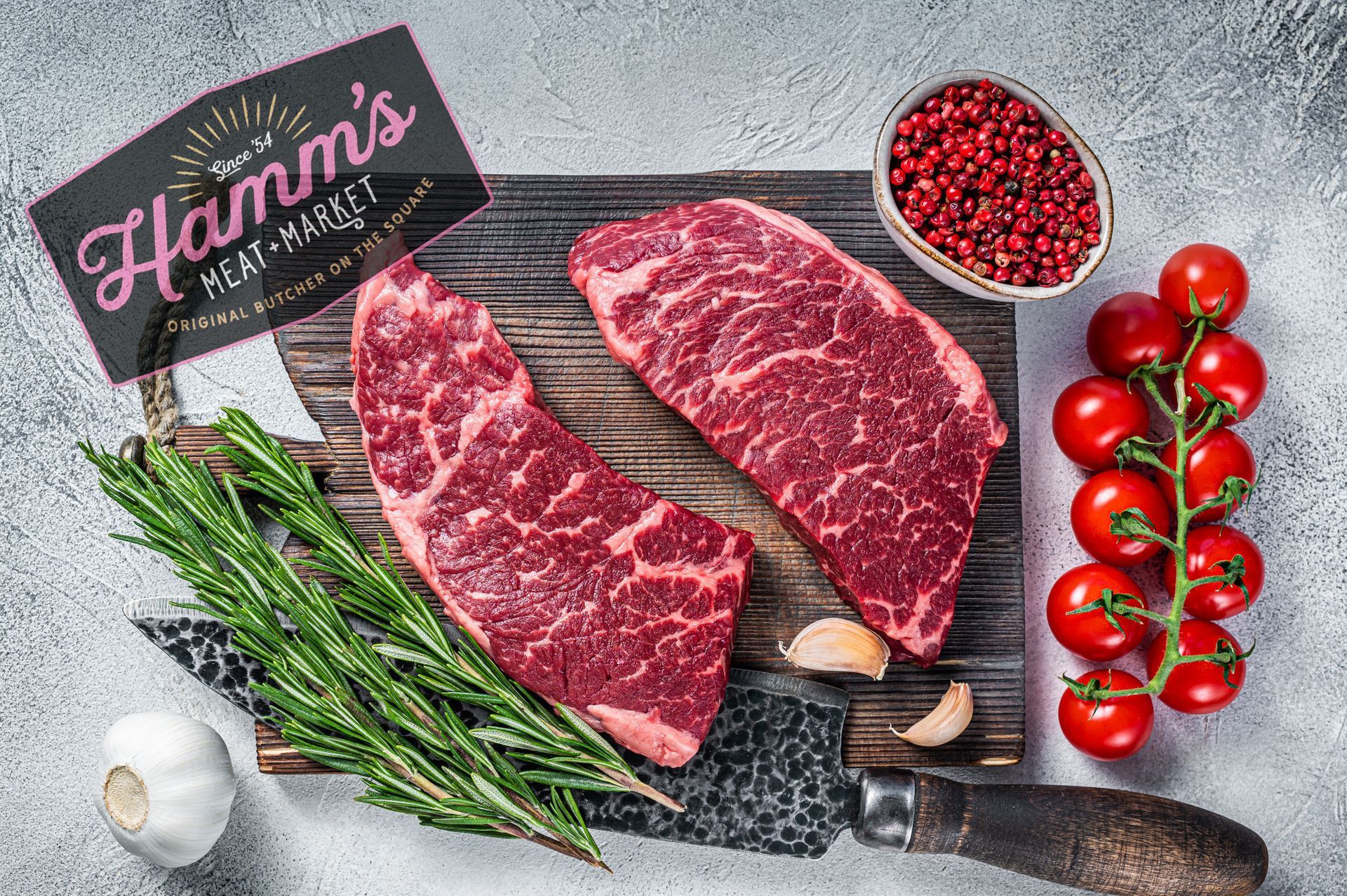 A cutting board topped with raw steaks , tomatoes , garlic and rosemary.