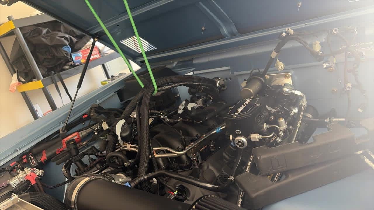 Open car engine bay with exposed black engine components and raised hood indoors