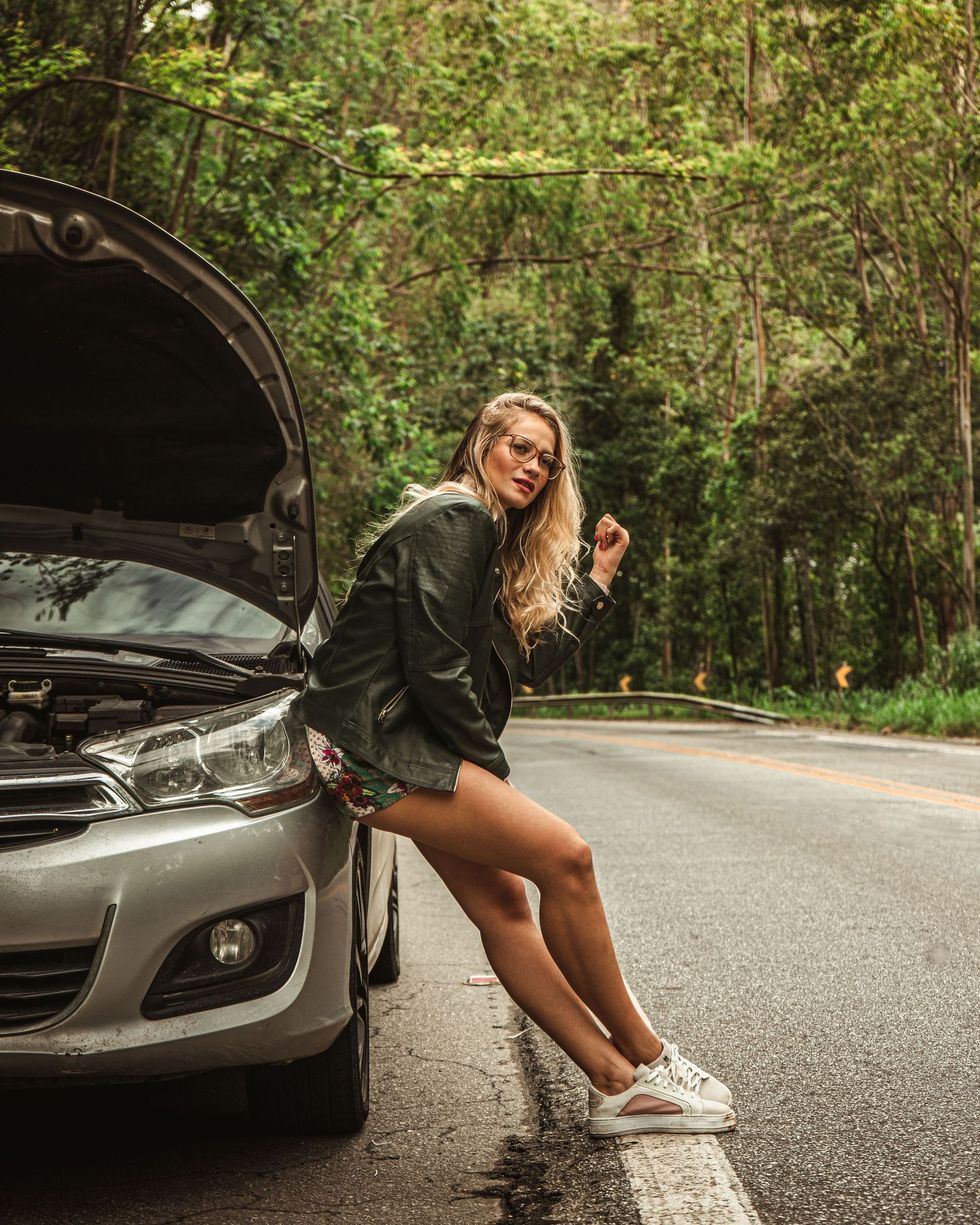 Woman sitting on a silver car with hood open on a forest road, holding a key fob.