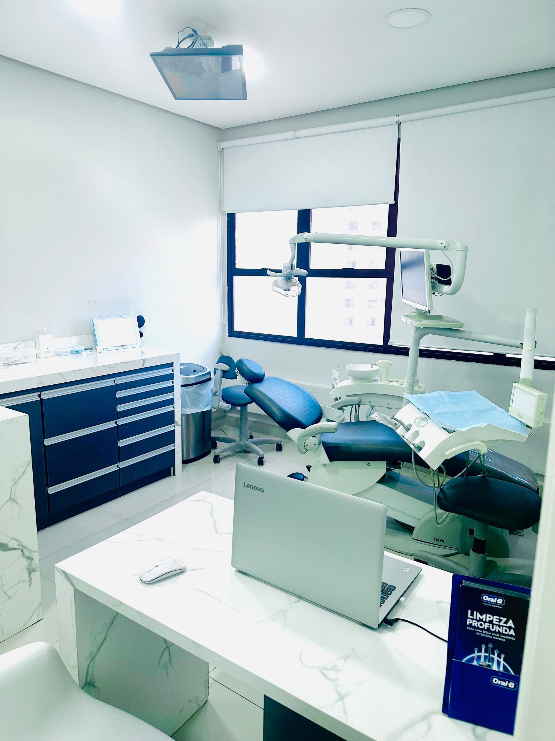 Sala de exames odontológicos com cadeira, equipamento, mesa e um laptop.