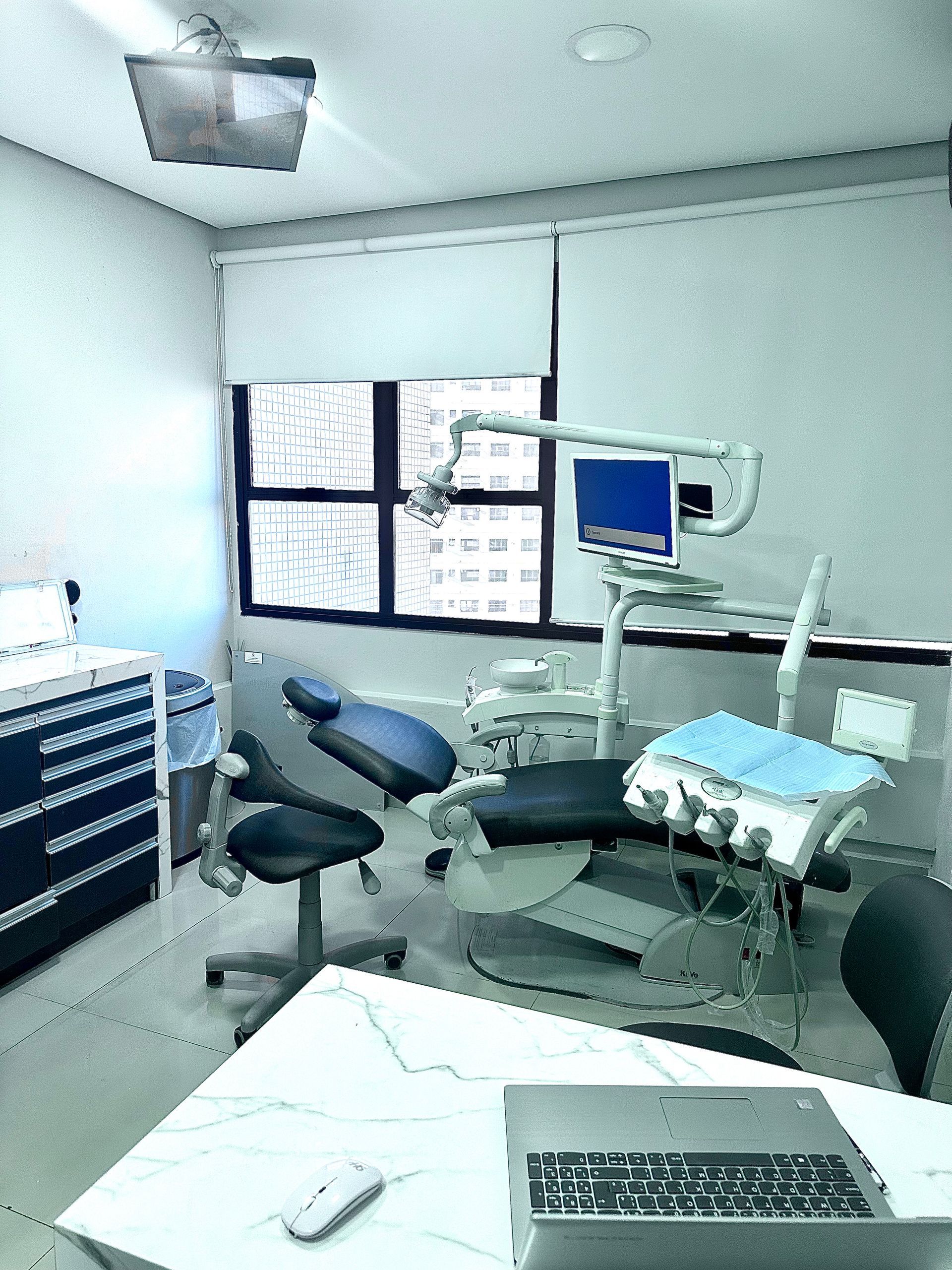 Consultório odontológico: cadeira odontológica azul, equipamentos, janela e computador.