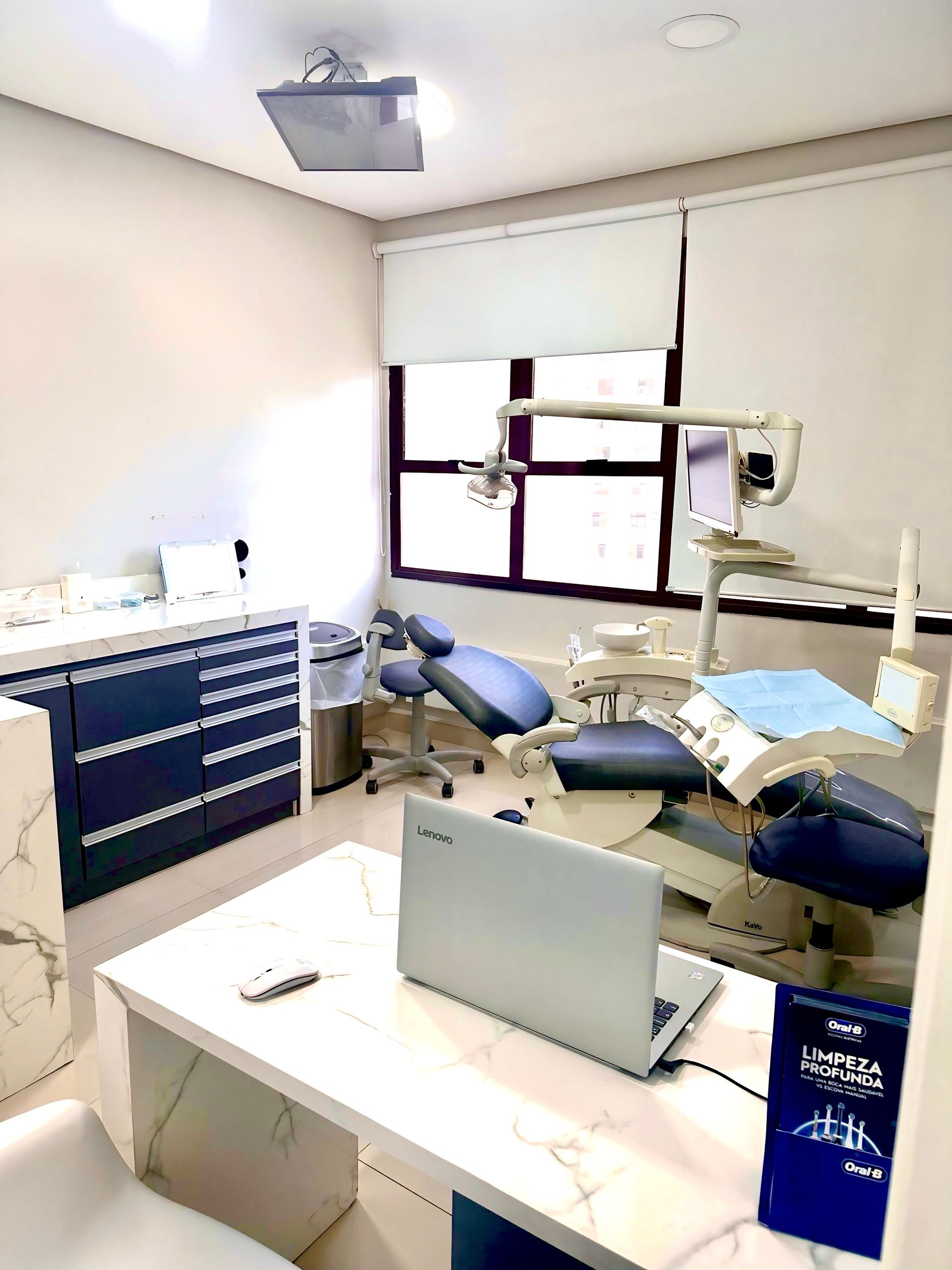 Consultório odontológico com cadeira odontológica azul, mesa e armários. Um laptop e uma janela são visíveis.