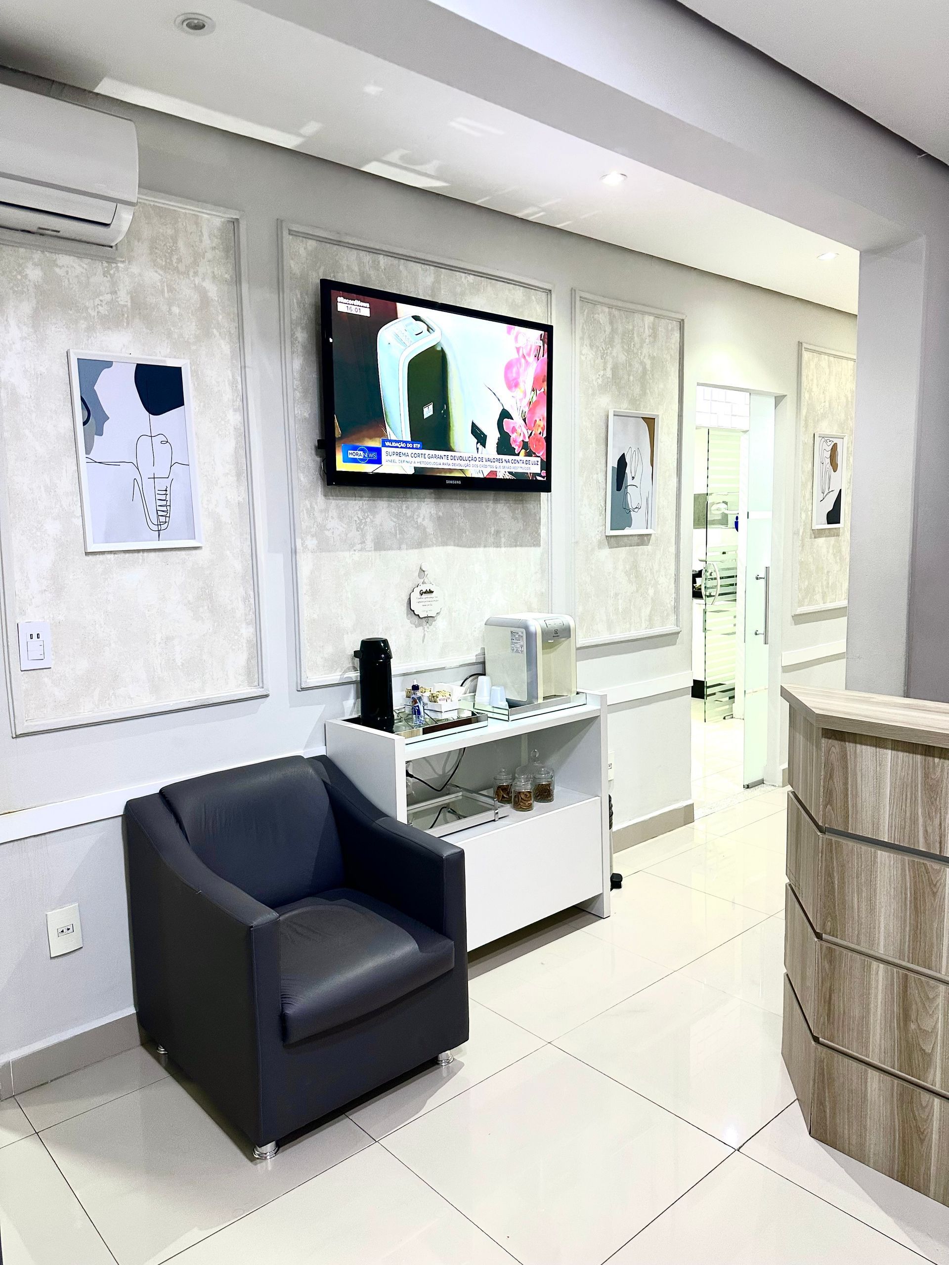 Sala de espera com cadeira cinza, armário branco, TV e quadros.