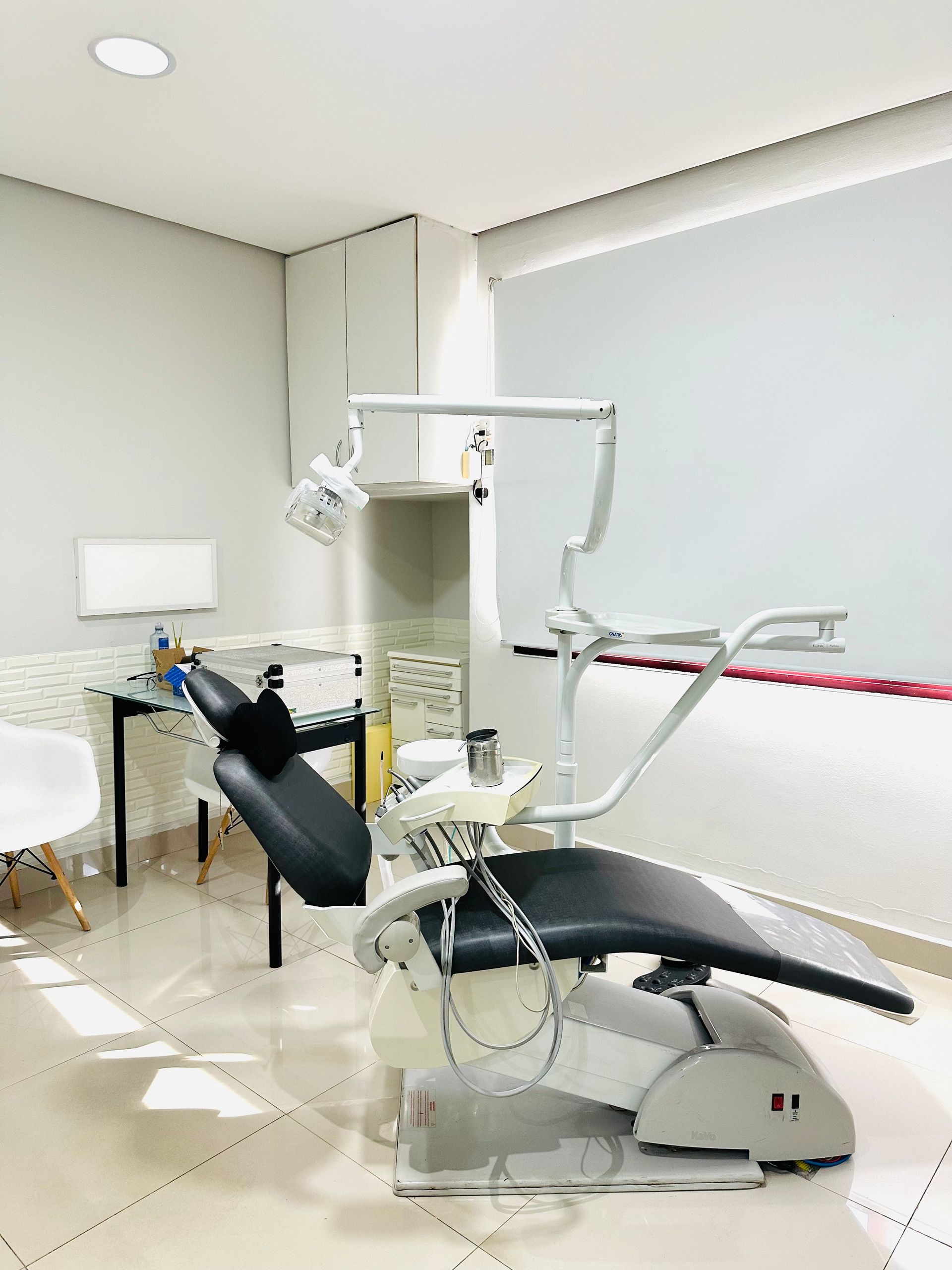 Consultório odontológico com cadeira odontológica preta, instrumentos, mesa e janela.