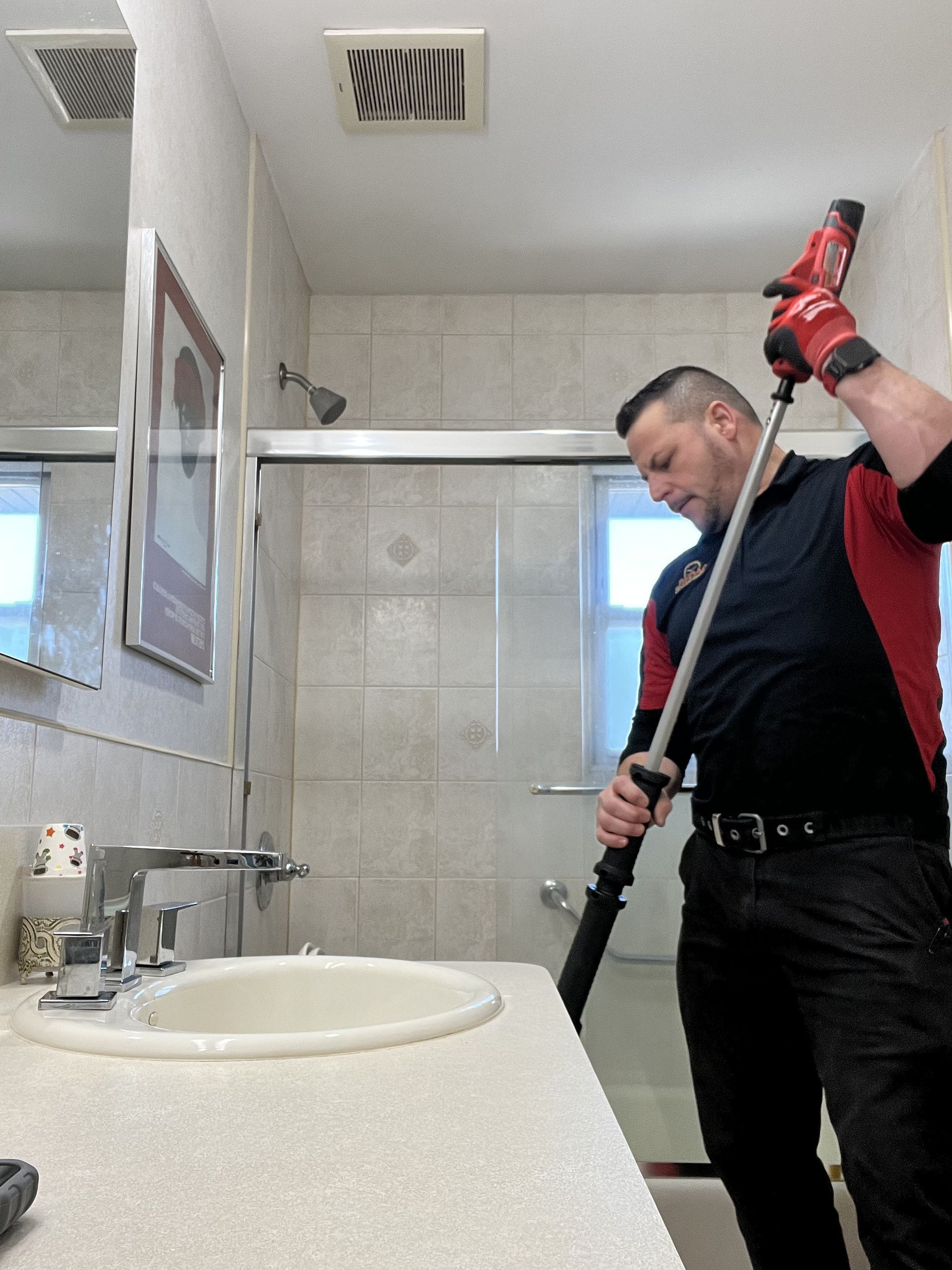 Man in bathroom using a tool to clean; wearing black and red work attire.
