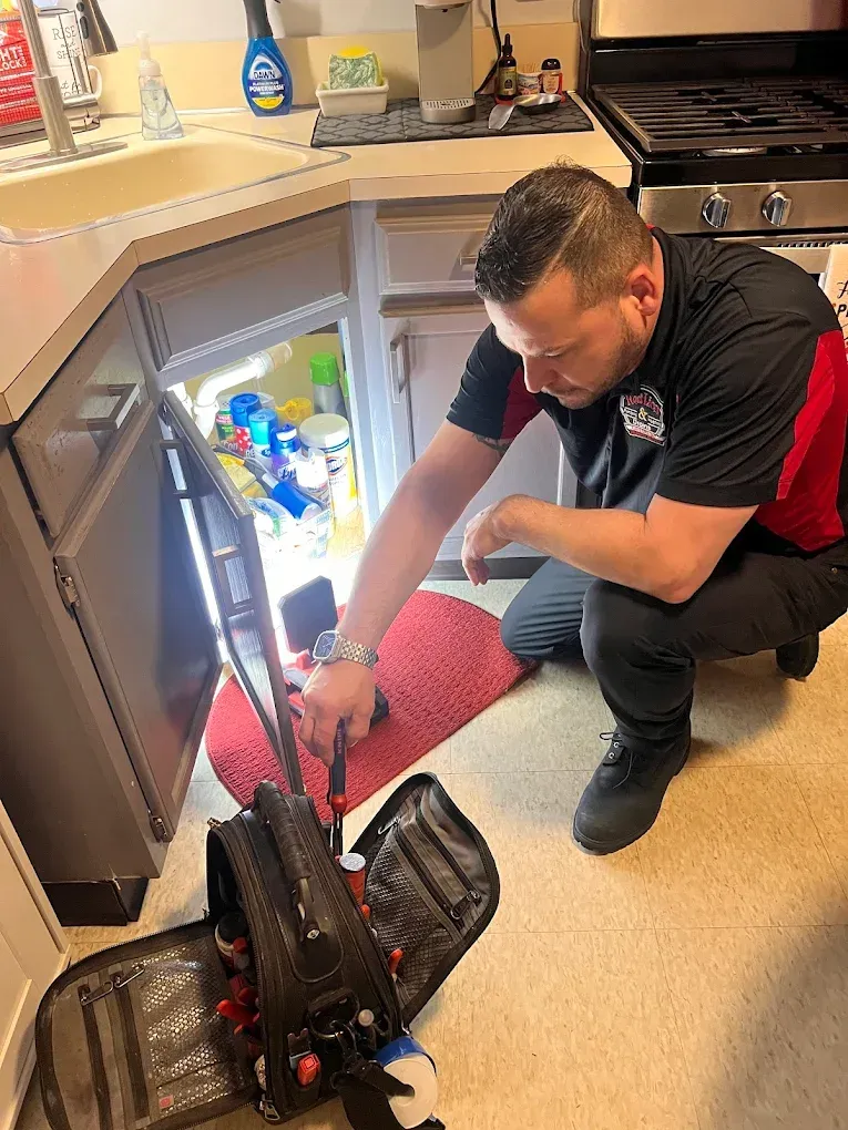 A man in black/red shirt repairs a kitchen appliance, tools at his side.