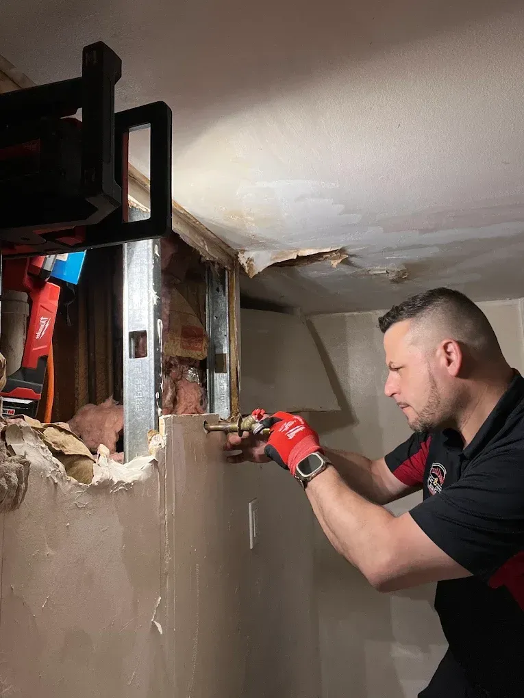 Man in red gloves working on plumbing inside a wall, in a basement.