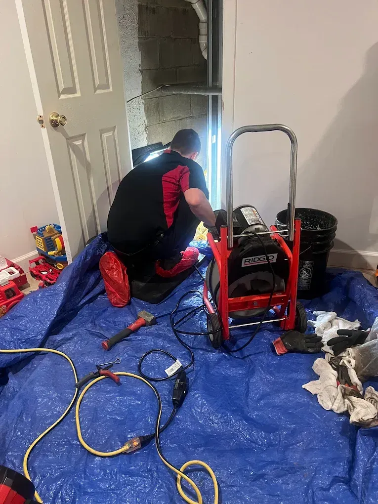 Plumber in red and black shirt working on a pipe, with equipment on blue tarp in a room.