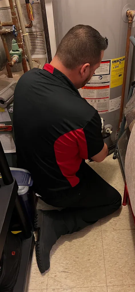 A person in black with red trim kneels to work on a water heater in a basement.