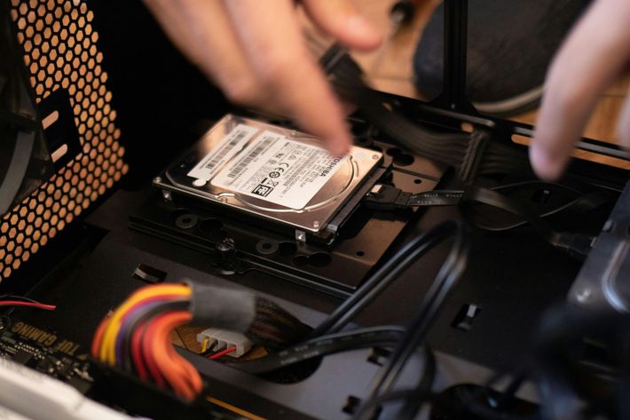 Person installing a hard drive into a computer case, pointing at the drive. Black and gray components are visible.