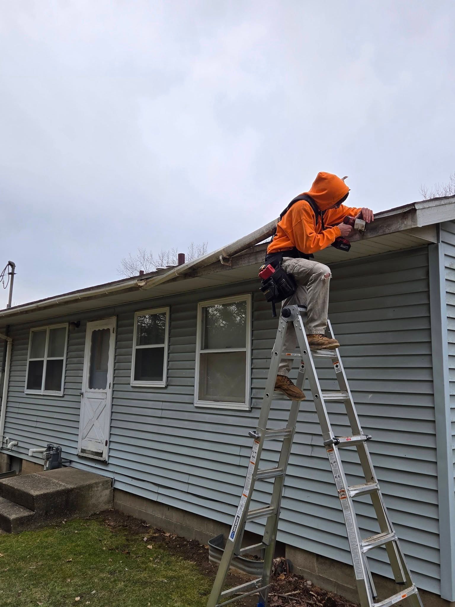 Worker in orange jacket on ladder, repairing a gray house roofline under an overcast sky.