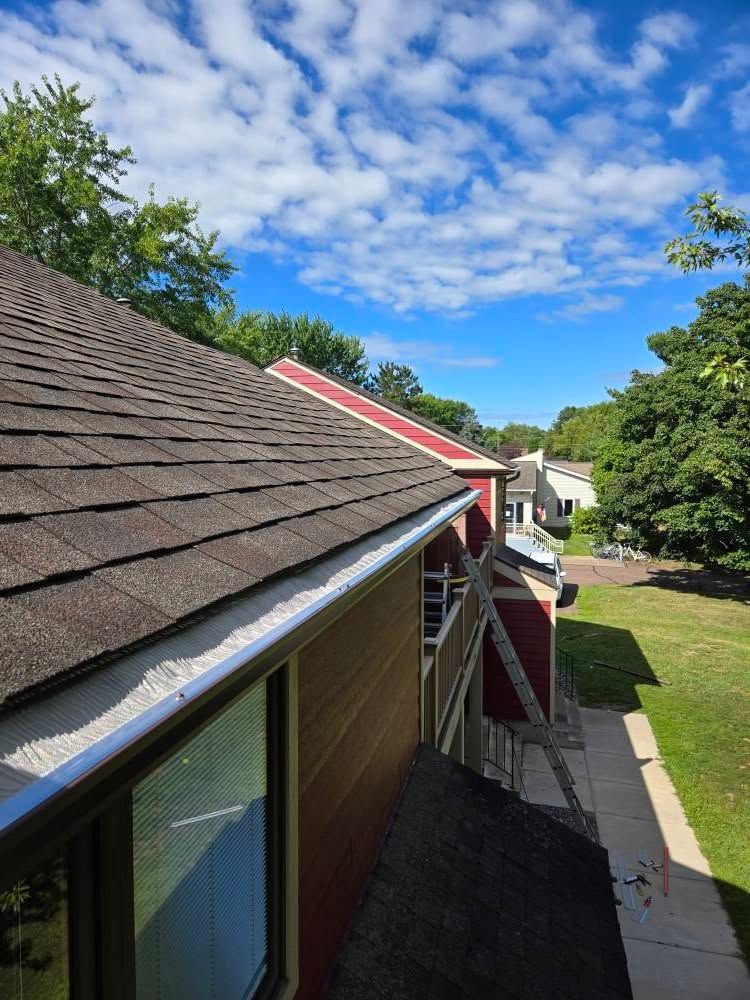 Roofline of a house with brown shingles, red trim, and blue sky with clouds above the trees