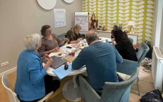People seated around a table, working. Light-filled room with round table and chairs. Some people are holding objects.