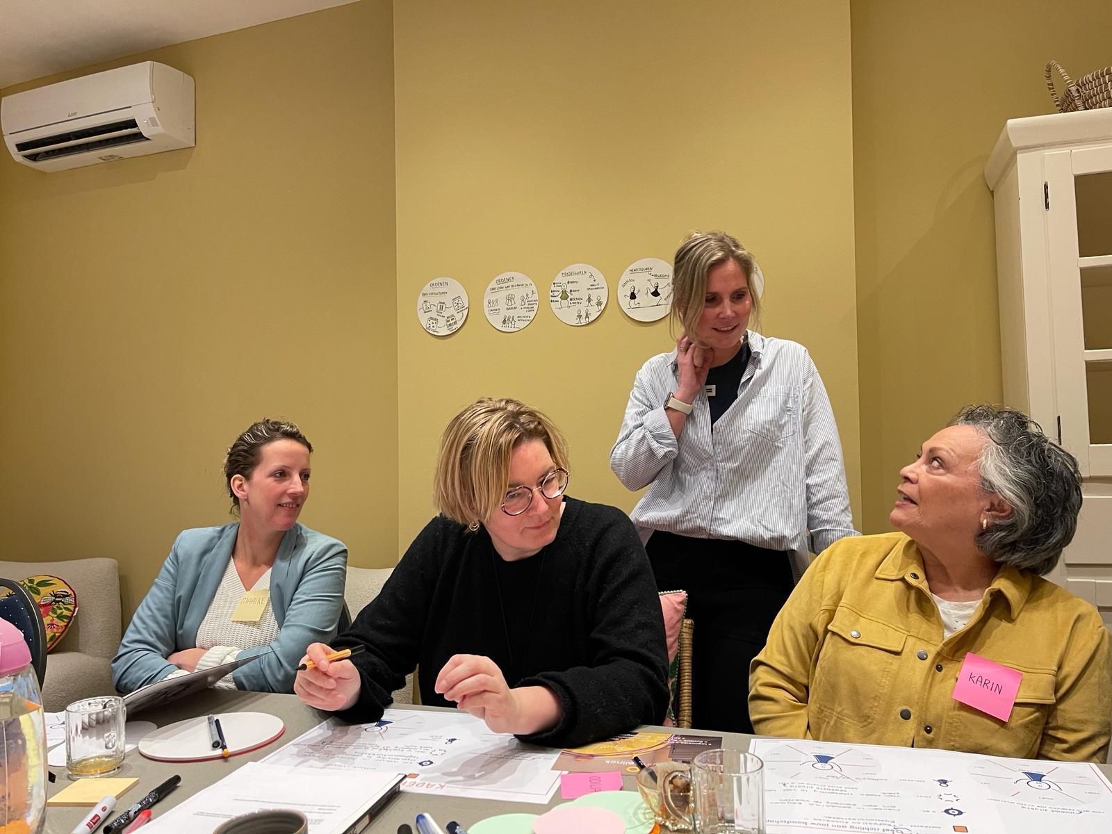 People seated around a table, working. Light-filled room with round table and chairs. Some people are holding objects.