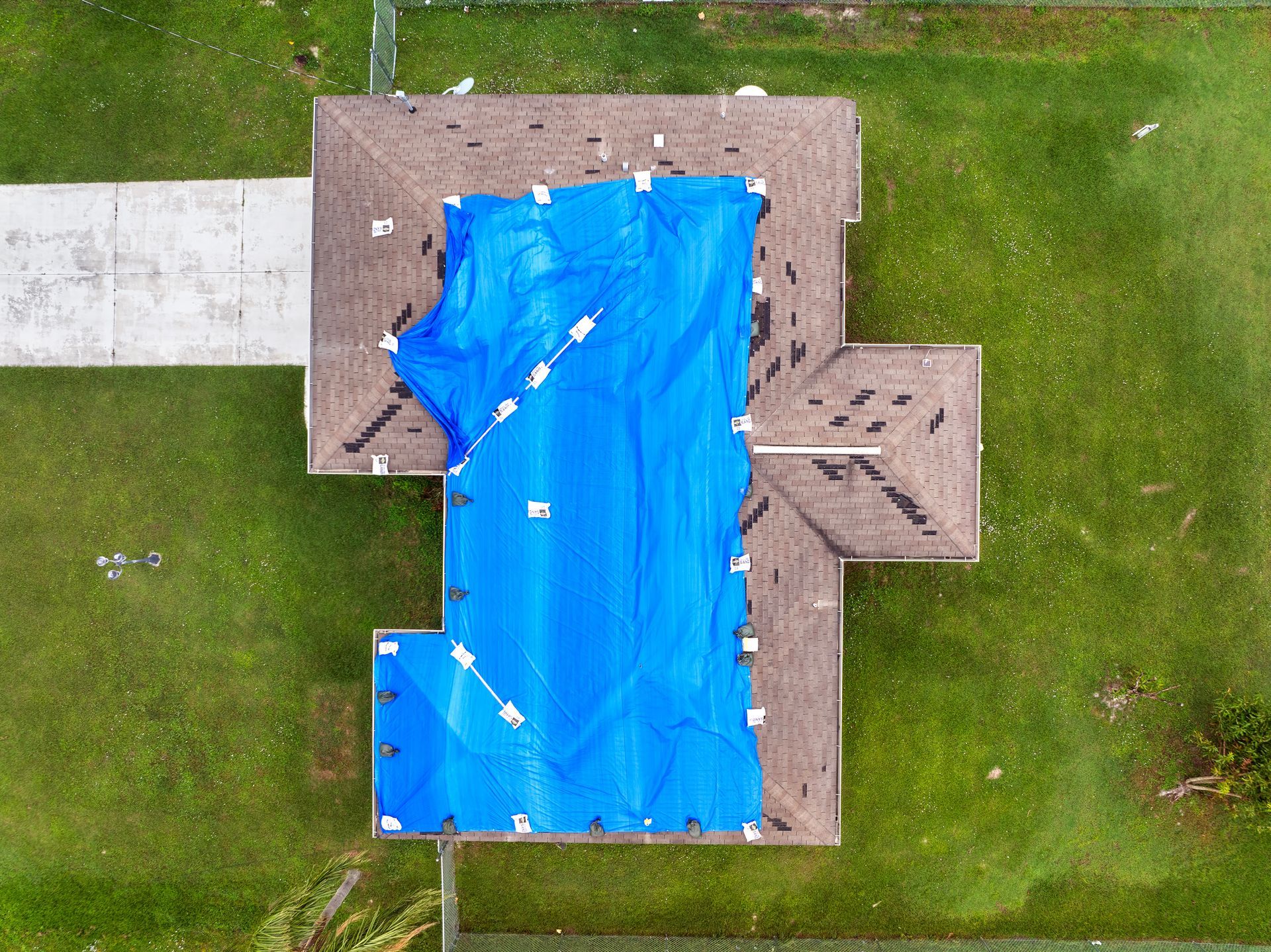 Overhead view of a house with a blue tarp covering a portion of the damaged brown roof. Green grass surrounds.